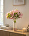 Eine Vase mit Alstroemeria rosa steht auf einem hölzernen Sideboard neben Büchern, einer Schale, einer brennenden Kerze und einer kleinen Skulptur. Sanftes Tageslicht fällt durch das Fenster und erfüllt den ruhigen Raum mit Frühlingsgefühlen.