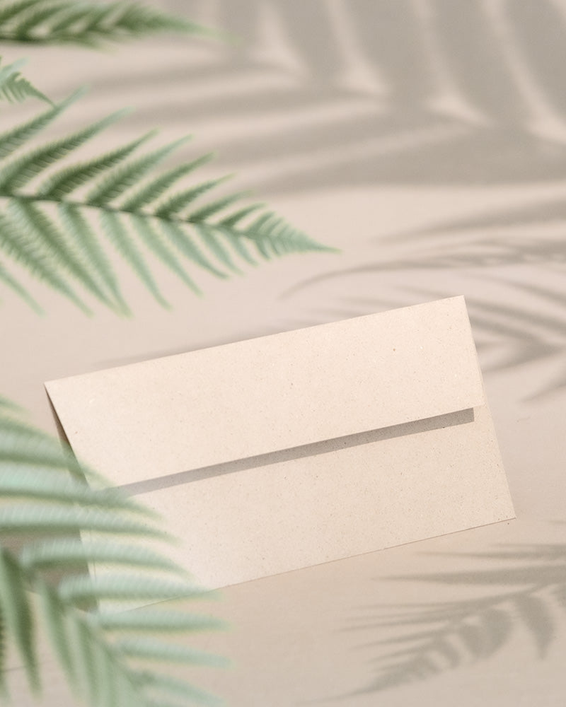 Ein Gutschein in Beige liegt aufrecht auf einer hellen Oberfläche, umgeben von sanften Schatten grüner Farnblätter und gleicht einem zarten Geschenk.