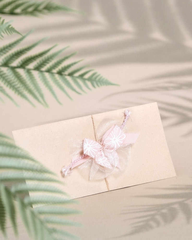 Ein beiger Gutschein, verschlossen mit einer rosa Schleife und geschmückt mit einem weißen Blumenmuster, liegt wie ein elegantes Geschenk auf einer Oberfläche, umgeben von zarten Farnschatten.