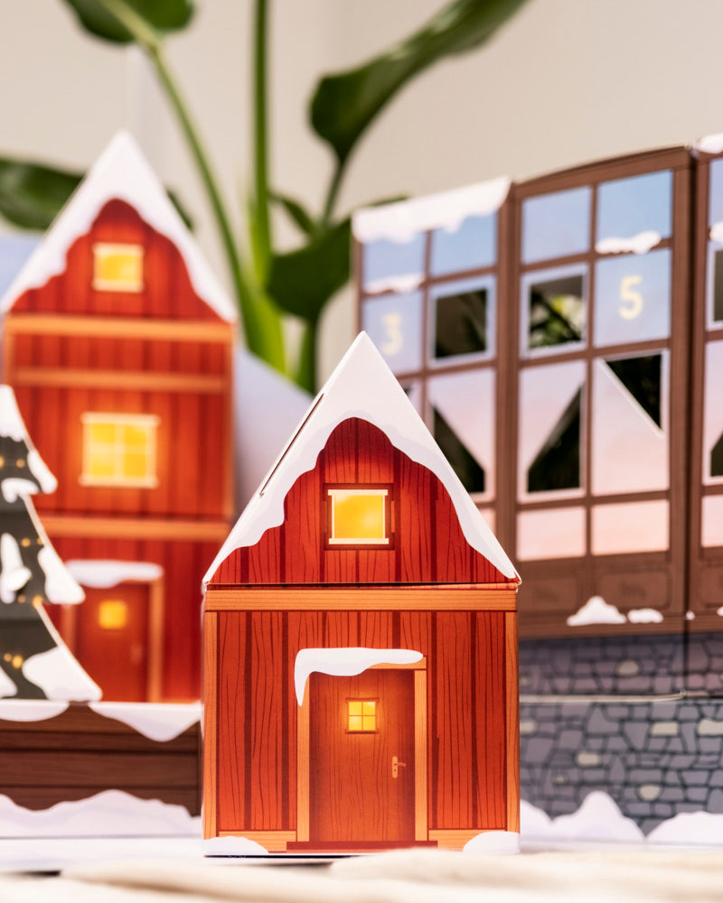 Ein Papiermodell eines verschneiten Holzhauses mit beleuchteten Fenstern steht vor festlichen Gebäuden und dem Pflanzen-Adventskalender mit echten Pflanzen und grünen Blättern im Hintergrund.