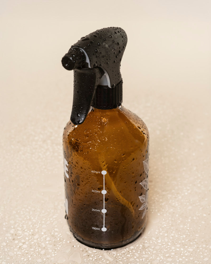 Ein brauner Pflanzensprüher mit schwarzer Auslösetülle steht aufrecht auf einer nassen, hellen Oberfläche, die mit Wassertropfen bedeckt ist. Auf der Vorderseite der Flasche sind weiße Messmarkierungen angebracht.