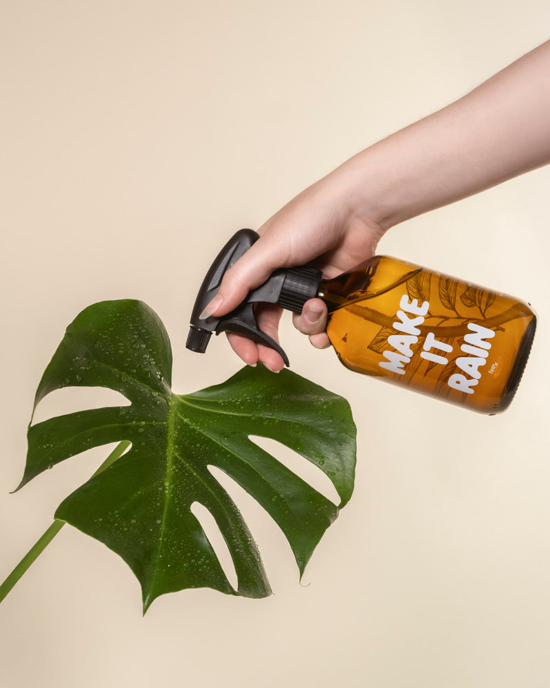 Eine Hand sprüht Wasser aus einem braunen Pflanzensprüher auf ein großes Monstera-Blatt, das sich vor einem hellbeigen Hintergrund befindet.
