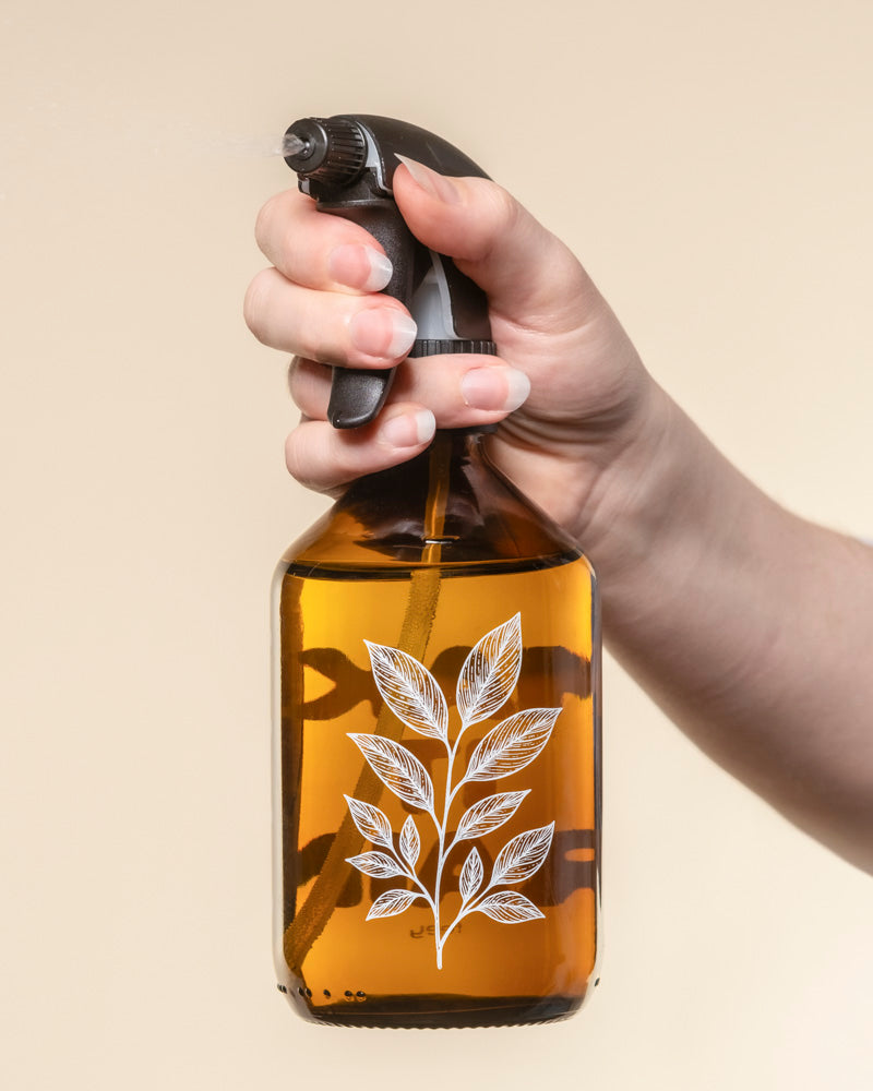 Eine Hand besprüht den braunen Pflanzensprüher mit einer schwarzen Düse, auf deren Vorderseite ein weißes Pflanzenblatt abgebildet ist. Vor einem beigen Hintergrund ist diese elegante Flasche perfekt zum Gießen Ihrer Lieblingspflanzen.