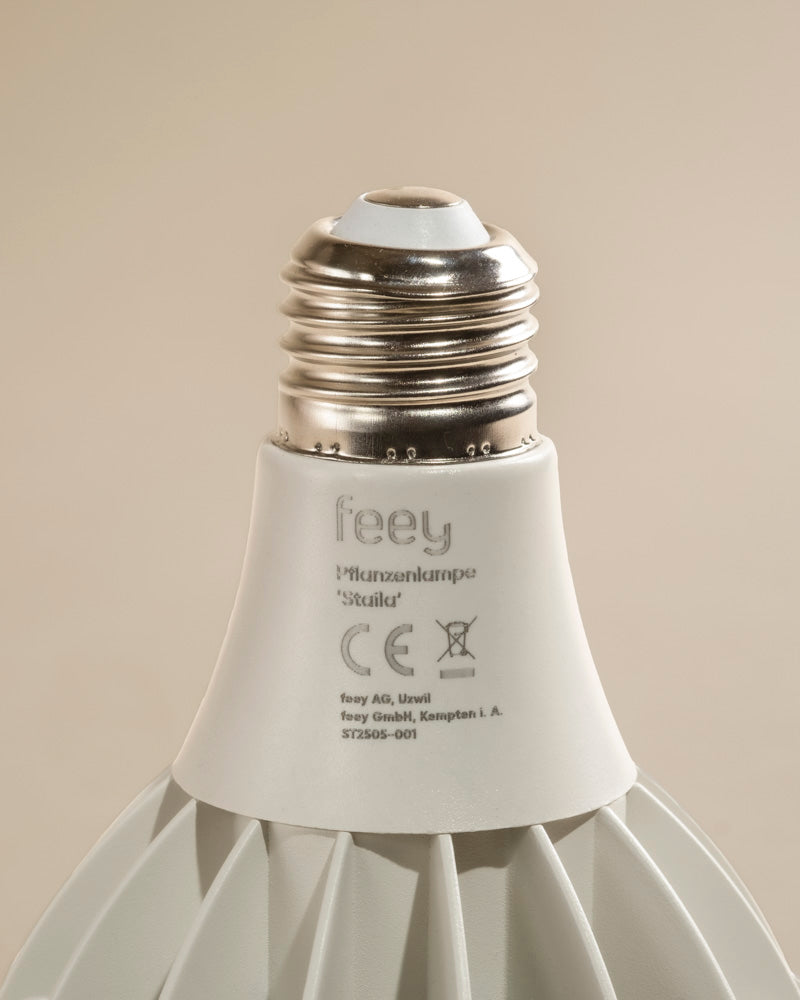Nahaufnahme des Sockels einer weißen Pflanzenlampe mit einer Metallverschraubung und einem Etikett mit Modellangaben und Zertifizierungssymbolen. Sie ist ideal für Zimmerpflanzen und bietet sonnenlichtähnliches Licht. Vor einem neutralen beigen Hintergrund.